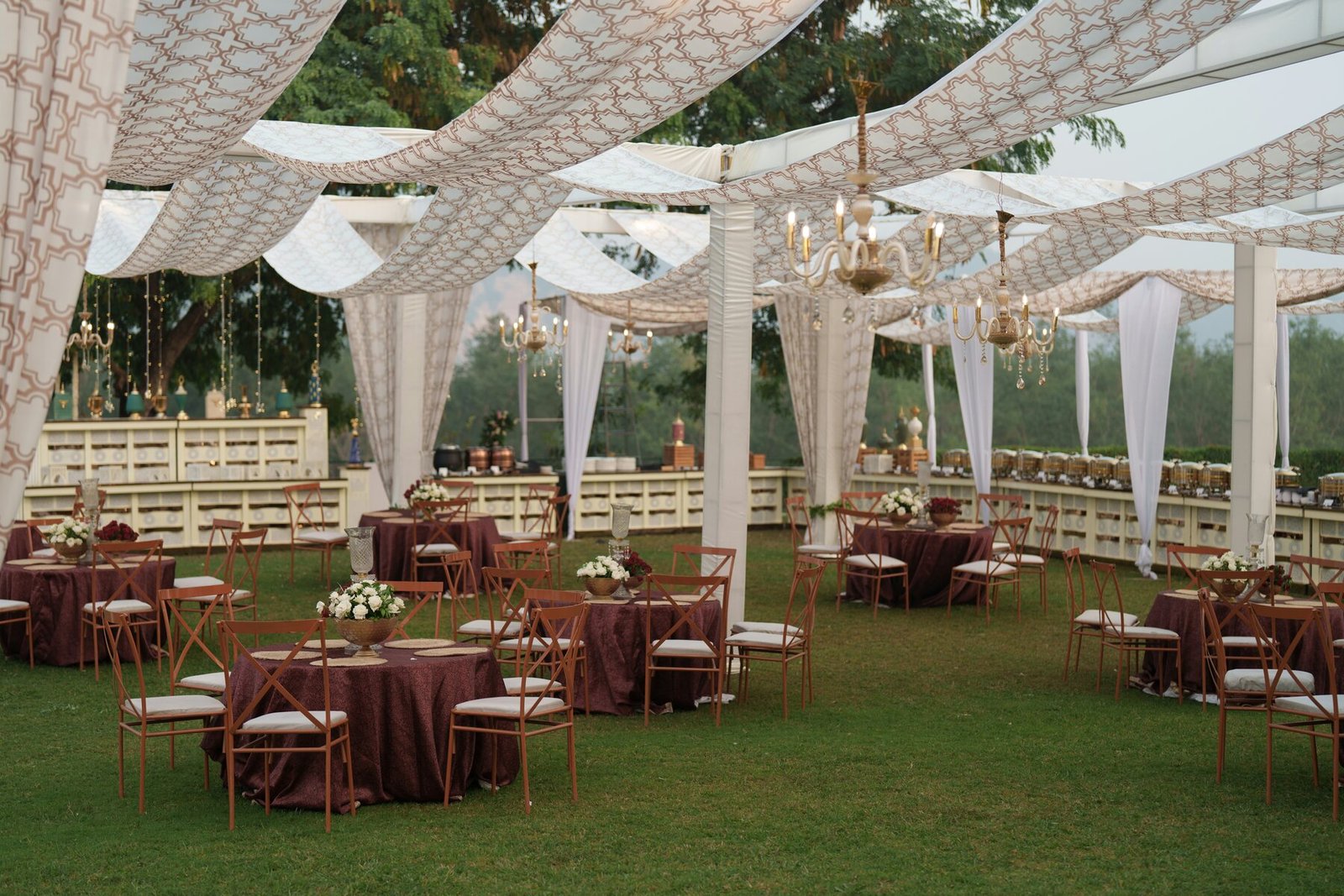 An elegant outdoor Indian wedding reception with draped canopies, banquet tables and a catered buffet arranged around the lawn